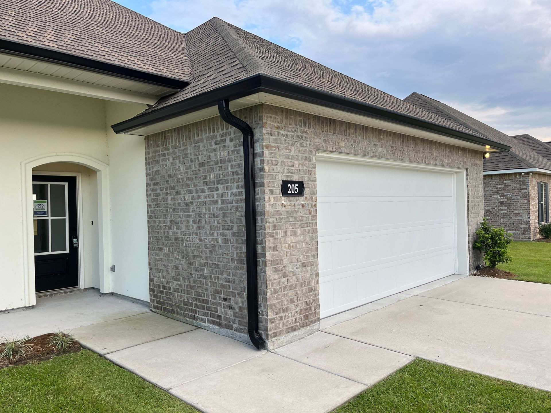 A residential house featuring a tan brick garage with a white door, black trim, and an adjacent white-walled entry.