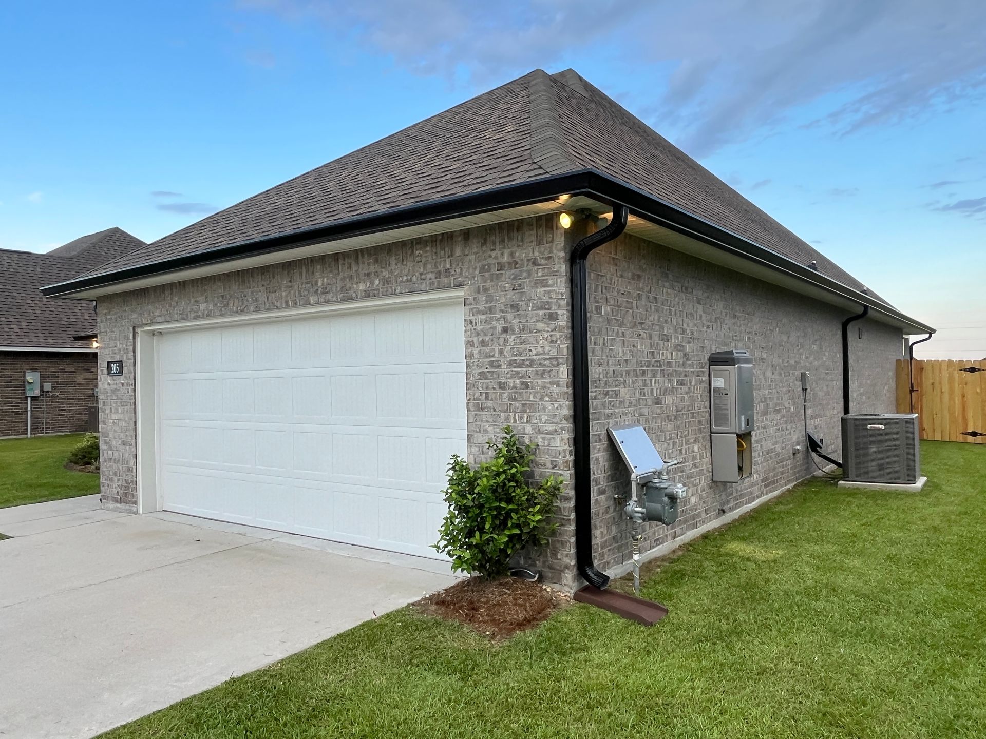 A gray brick garage with a white door and dark trim, featuring an AC unit and electrical box on the side yard.