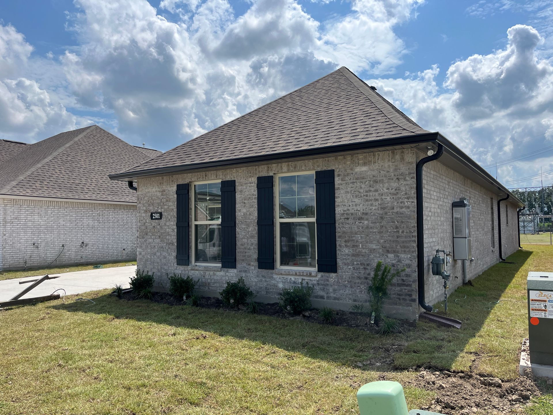 A side view of a modern, single-story light brick house with dark shutters, dark roof tiles, and a grass lawn under a sky.