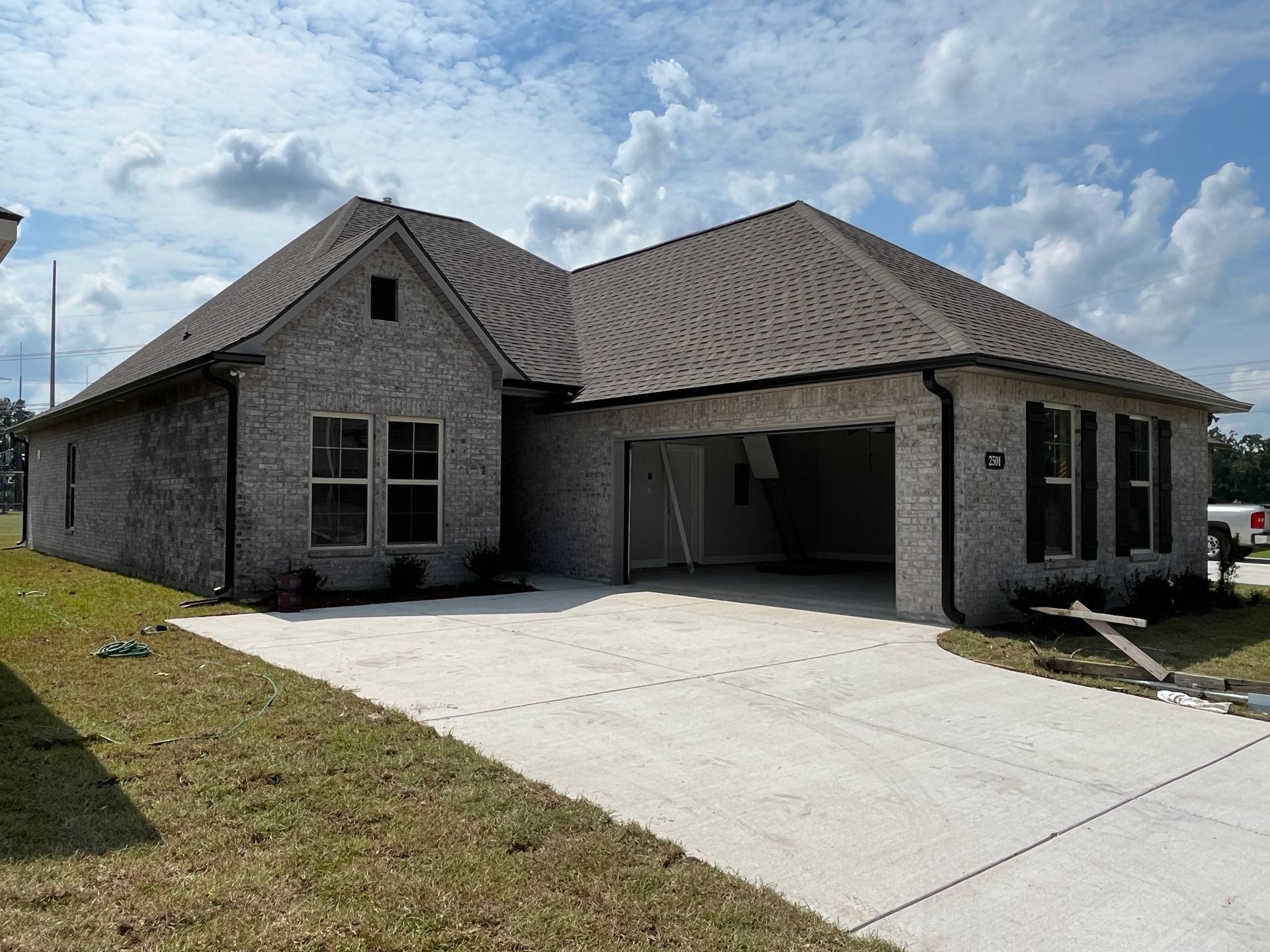 A newly built single-story suburban house with light gray brick, a dark shingled roof, and an open garage, seen from a lawn.