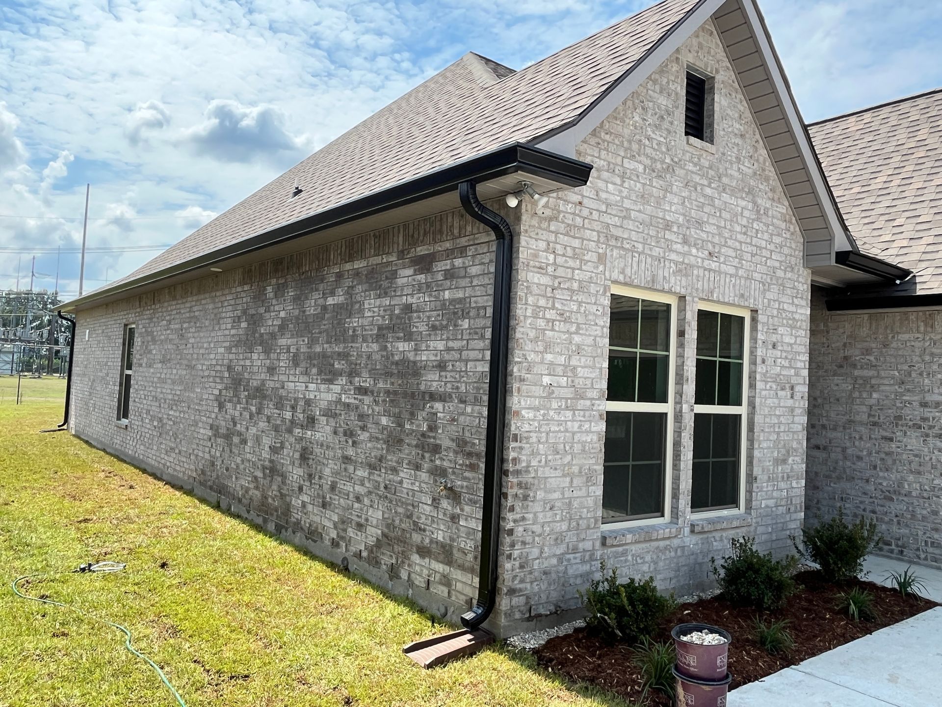 A single-story, light-colored brick house exterior with a gable roof, black gutters, and two windows in a grassy yard.