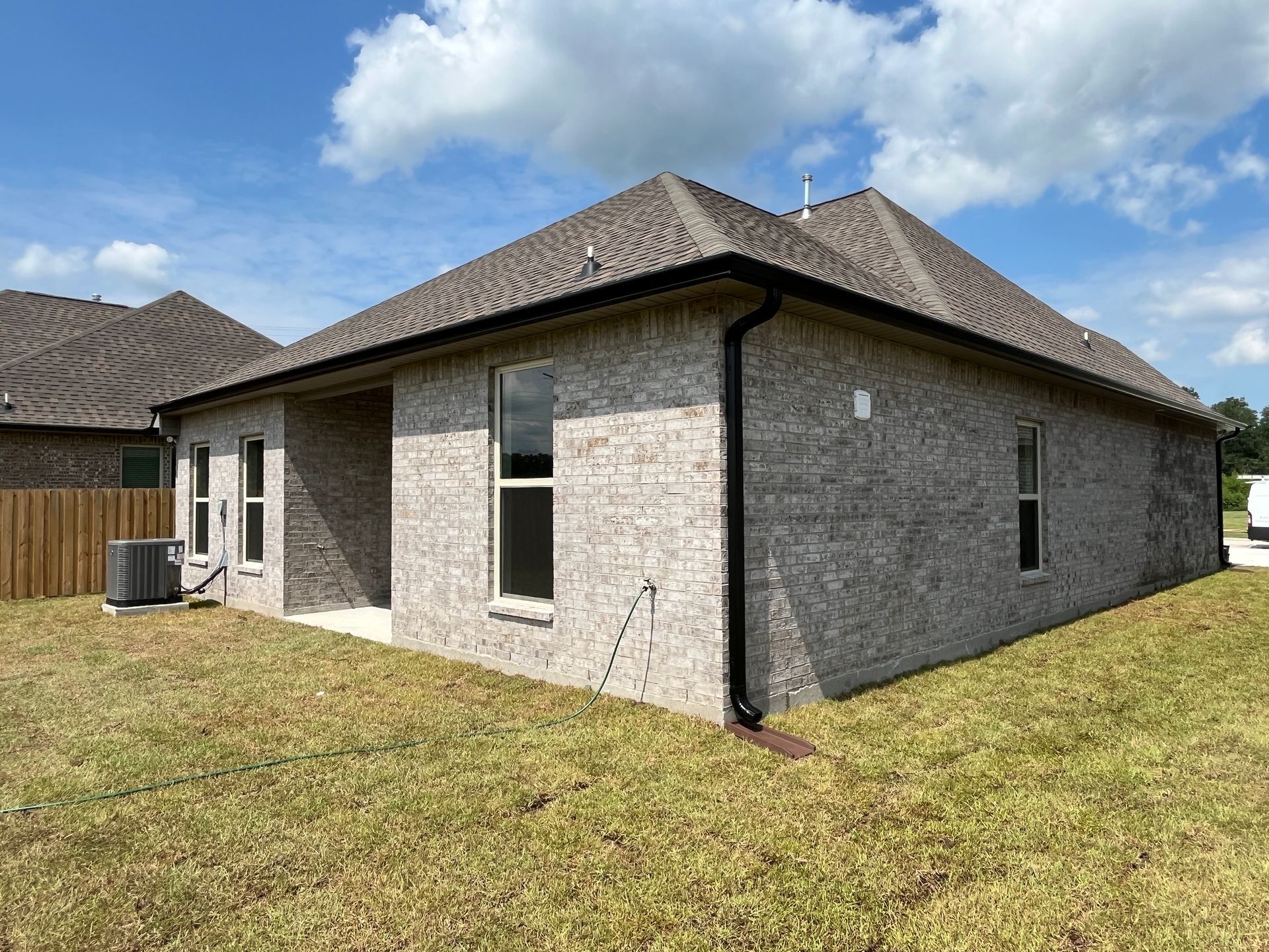 A single-story gray brick house with a dark shingle roof and a fenced yard under a sunny blue sky.