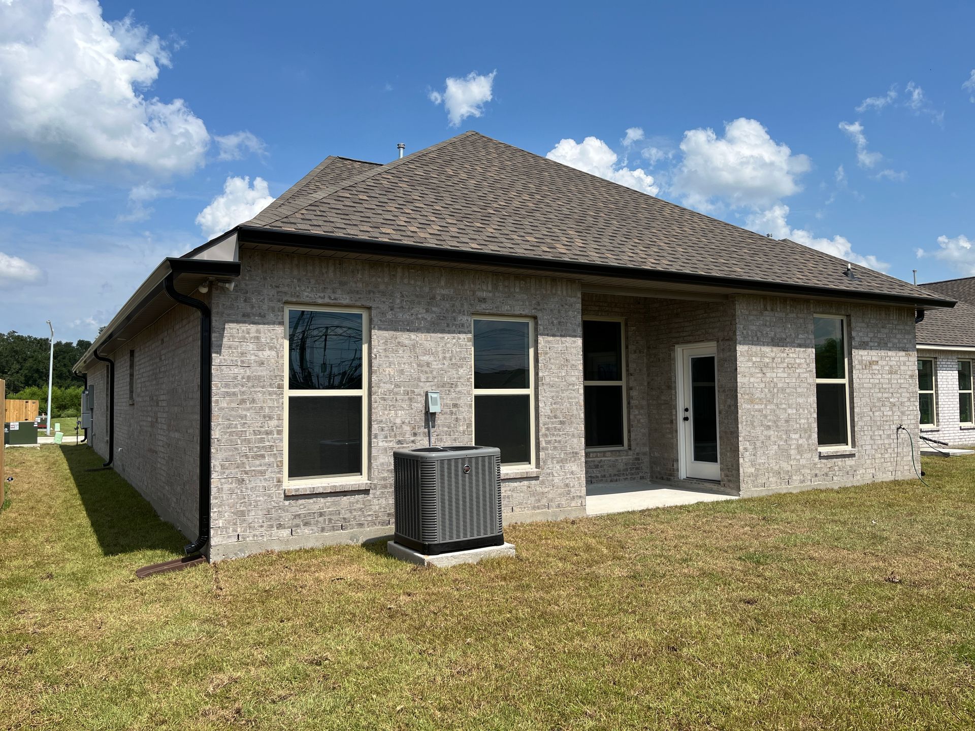 A gray brick single-story house with a shingled roof, lawn, and AC unit, seen from the backyard on a sunny day.