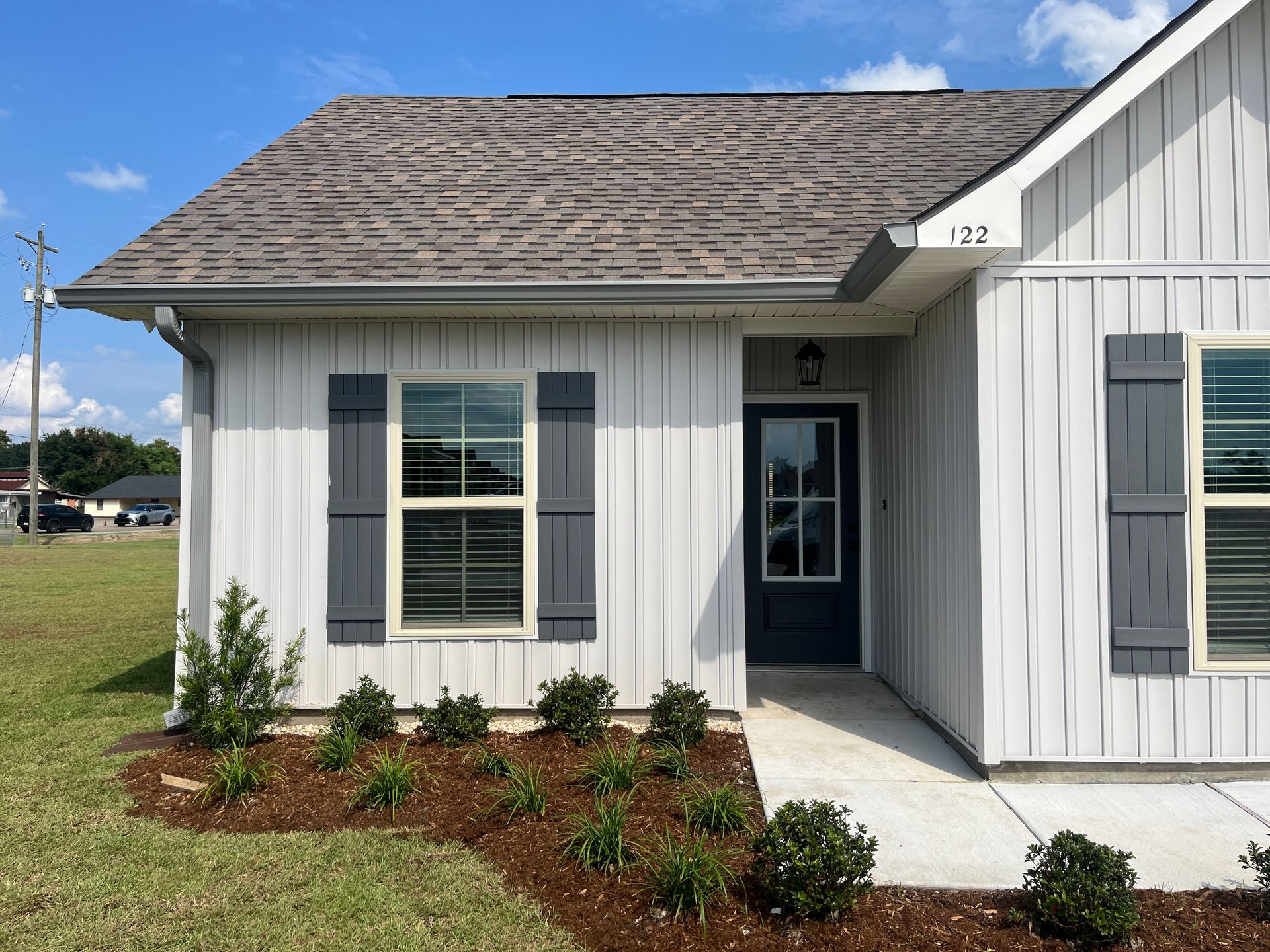 A one-story white house with vertical siding, a dark roof, gray shutters, and a front garden bed with small shrubs.