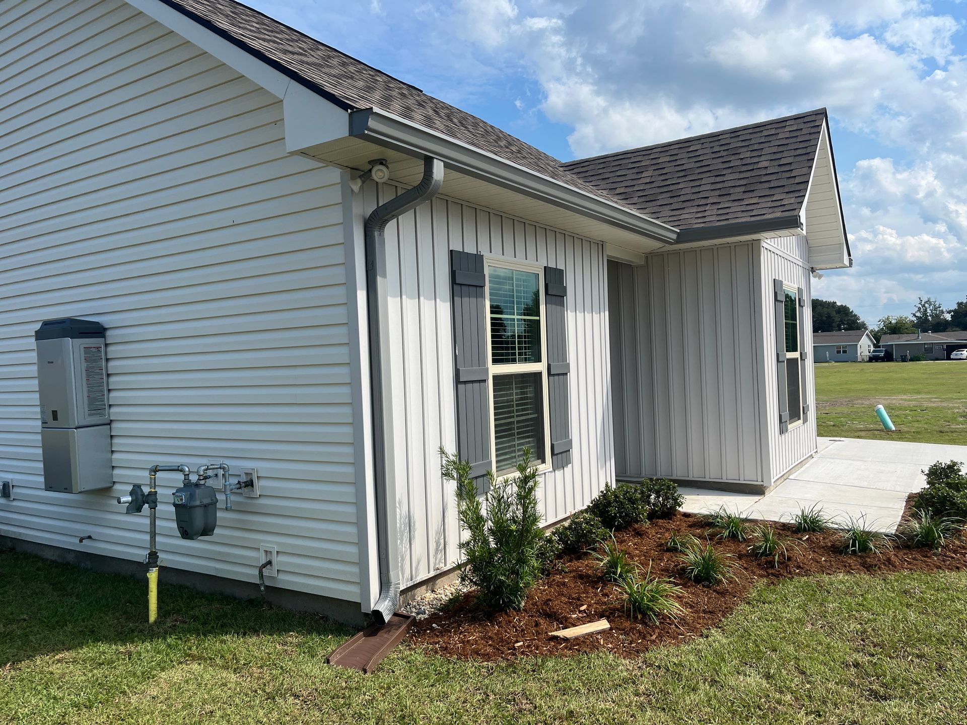 Side view of a white, single-story house with a grey roof, shutters, a utility meter, and fresh mulch landscaping.