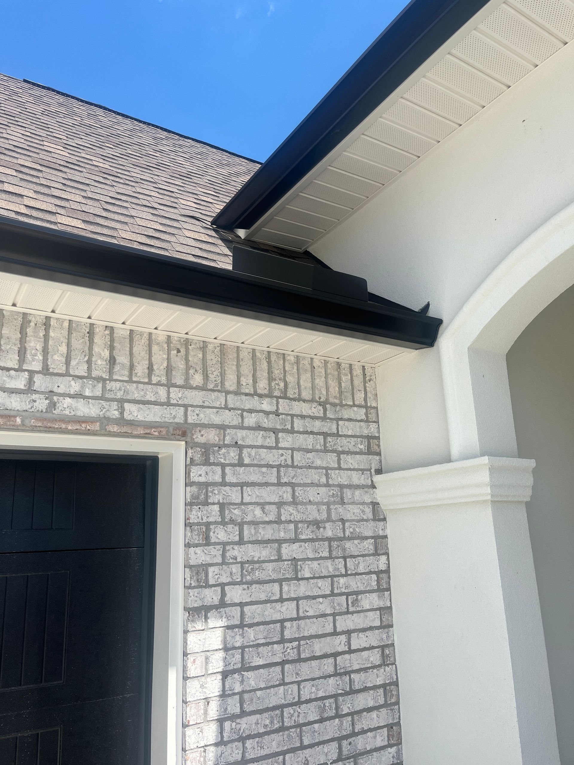 Close-up of a house corner showing a grey brick wall, black gutter, light-colored soffit, and a white arched entryway.