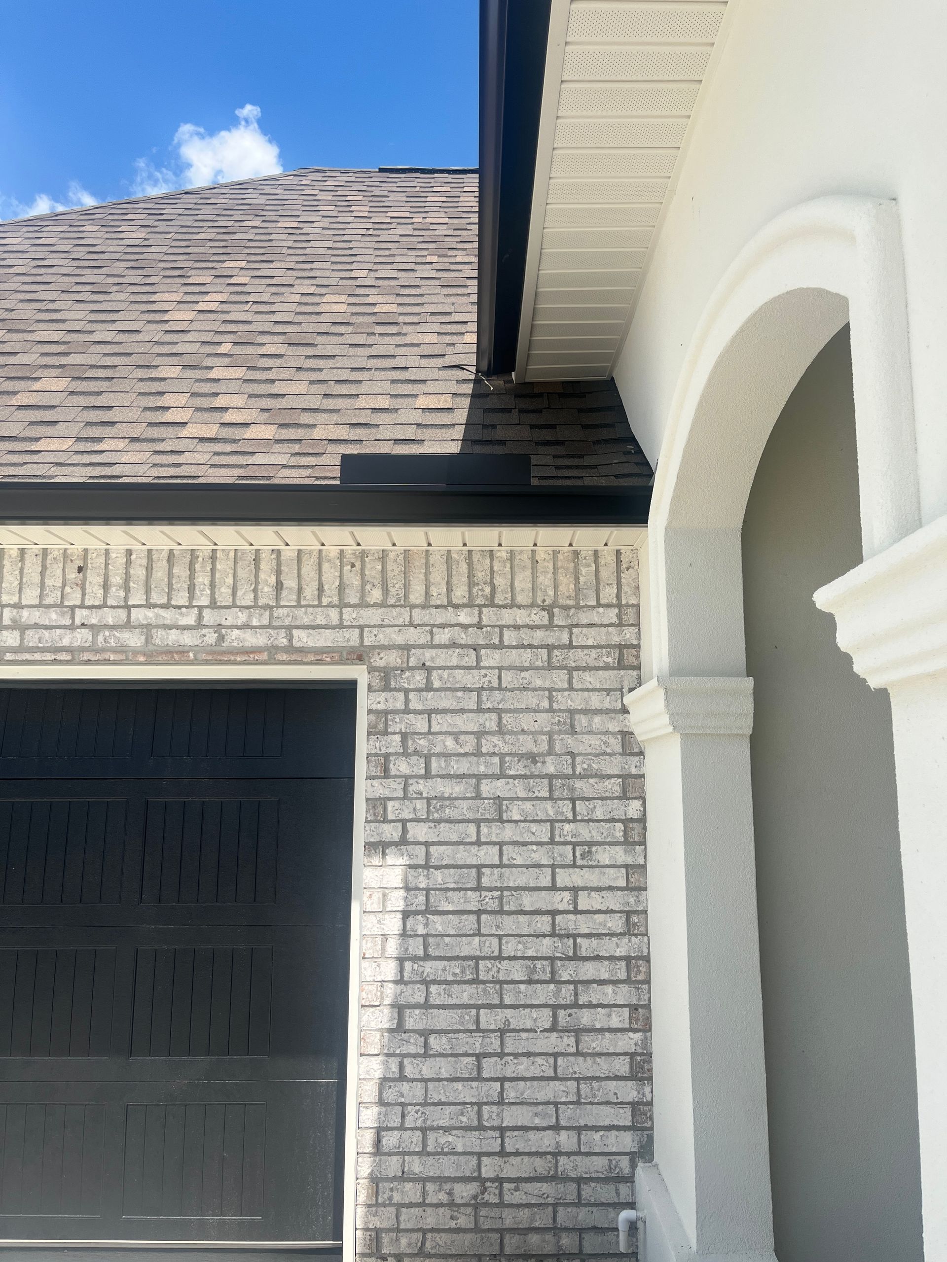A close-up of a house exterior showing a grey brick wall, a black garage door, a dark roof, and a white arched entryway.