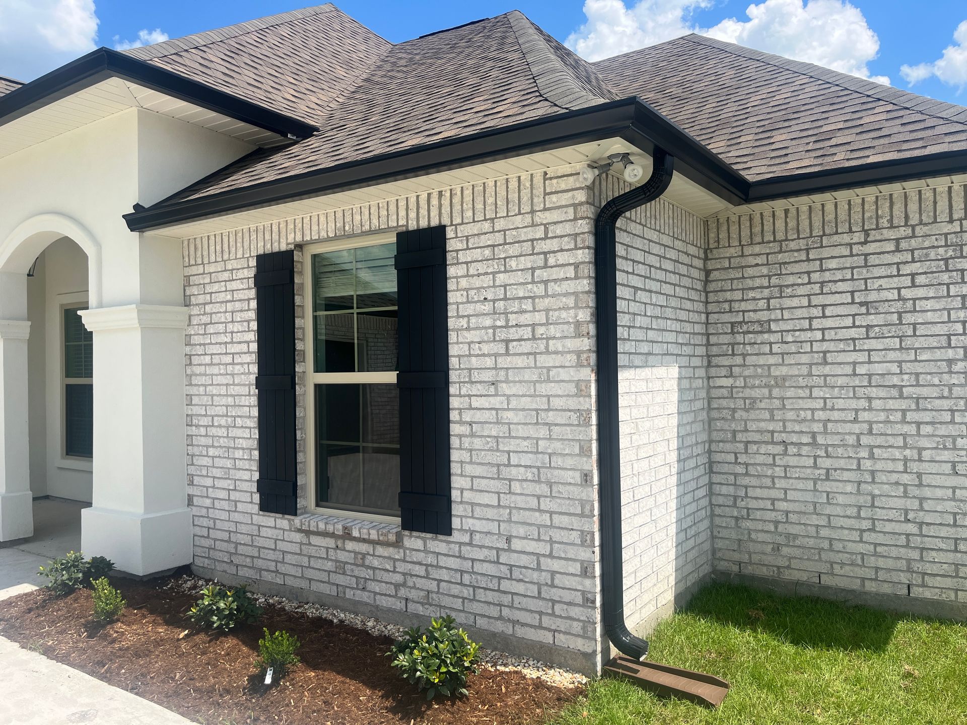 A one-story suburban home exterior featuring whitewashed brick, black shutters, a dark shingled roof, and green lawn.