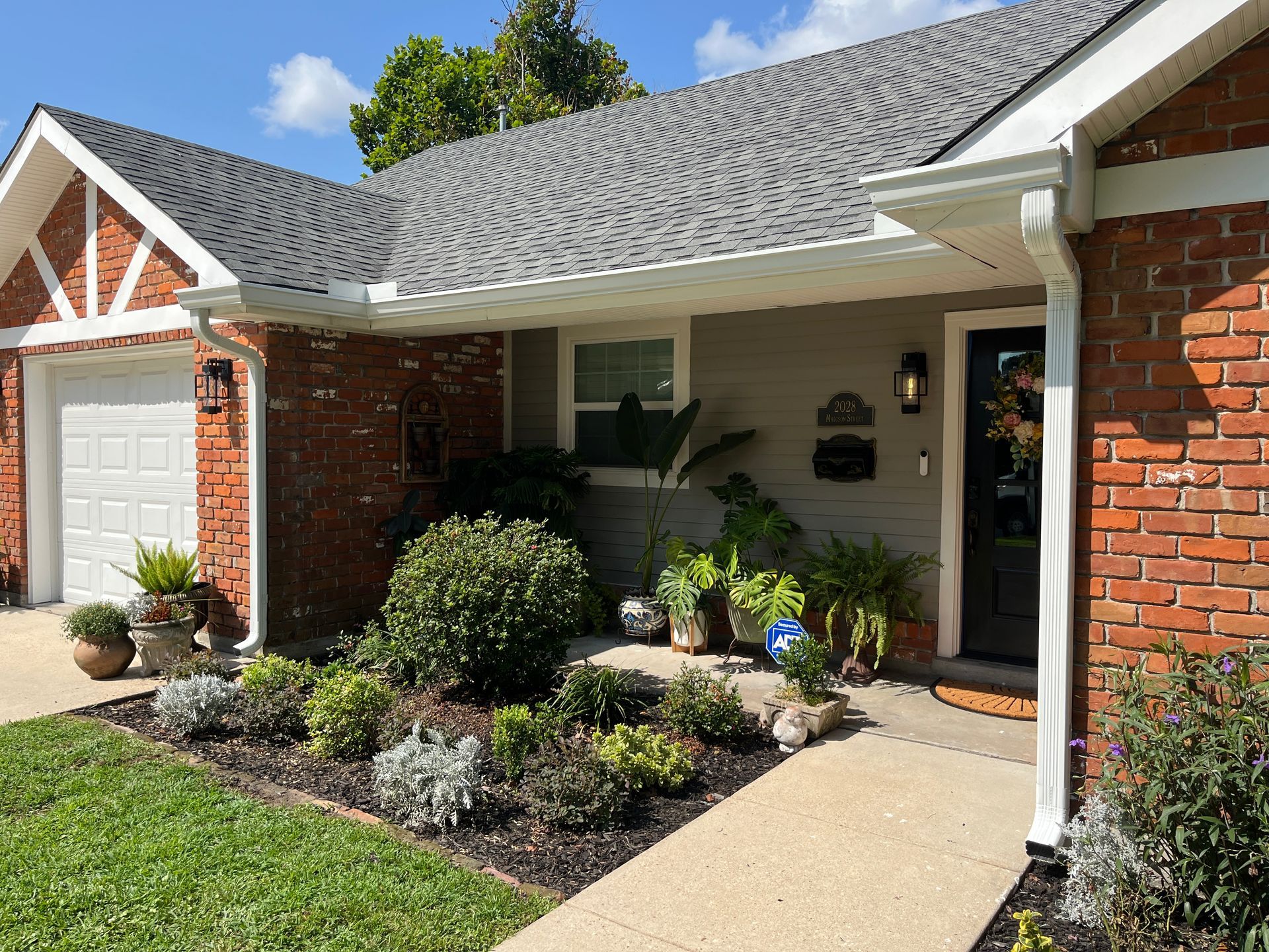 A brick-front suburban house with a gray shingled roof, a front porch, a garden, and a concrete walkway.