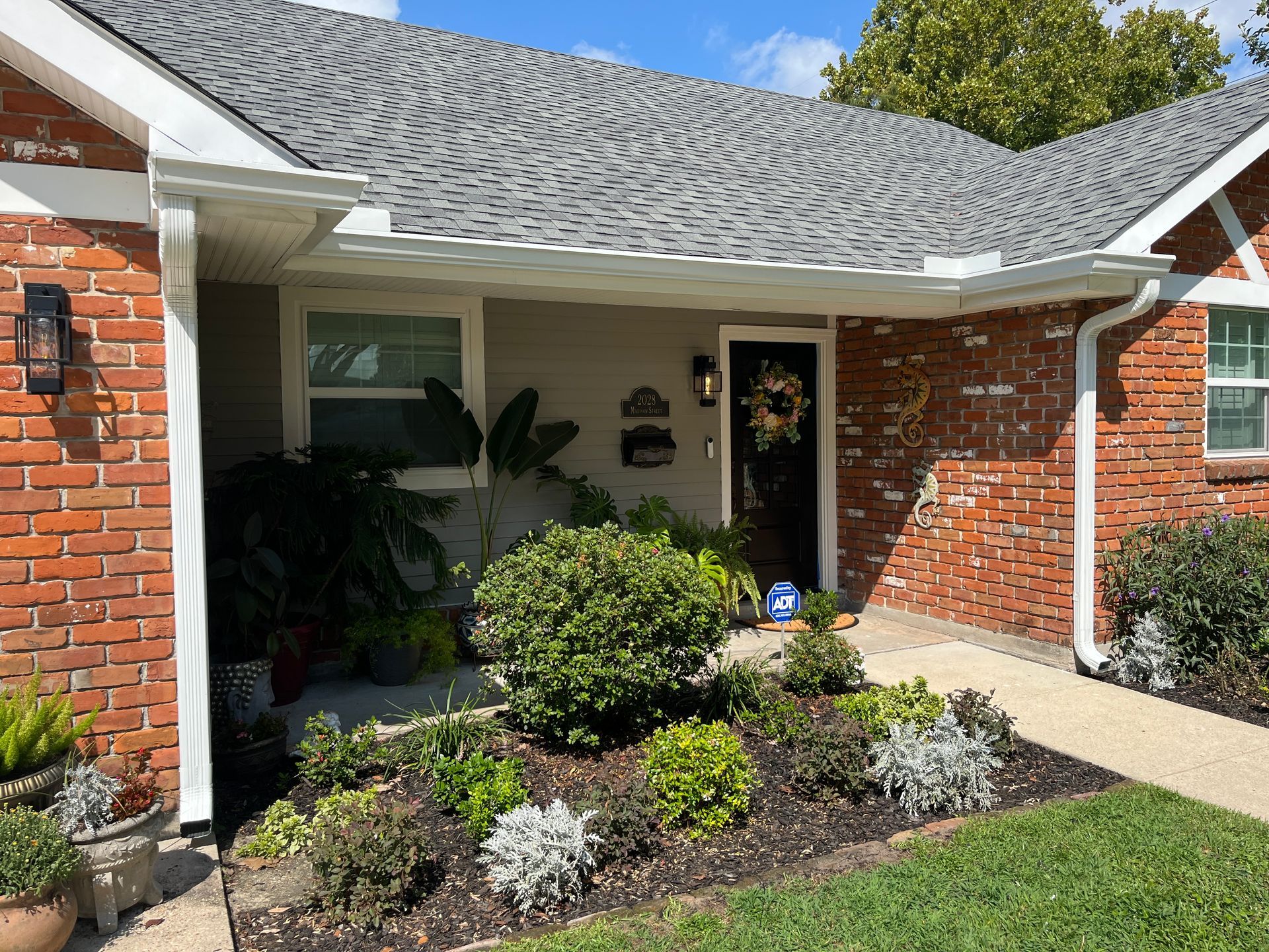A brick house with a front porch, a dark front door with a wreath, and a landscaped flower bed with various shrubs.