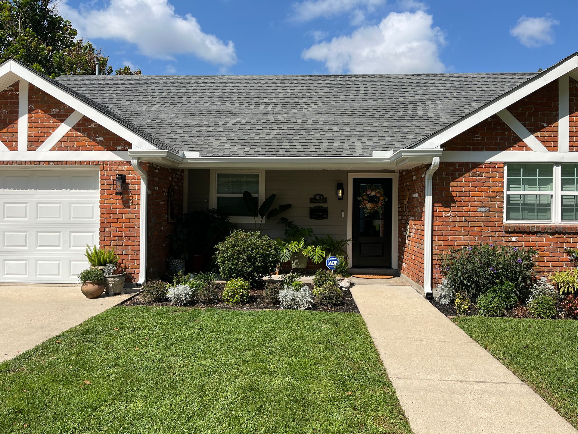 Single-story brick house with a white garage, gray shingle roof, front walkway, and green lawn under a sunny blue sky.