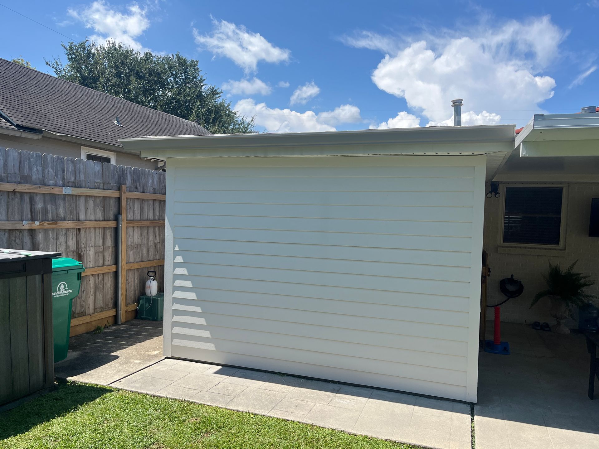 A white, horizontal-sided storage shed sits on a concrete patio in a yard with a wooden fence and blue, cloudy sky.