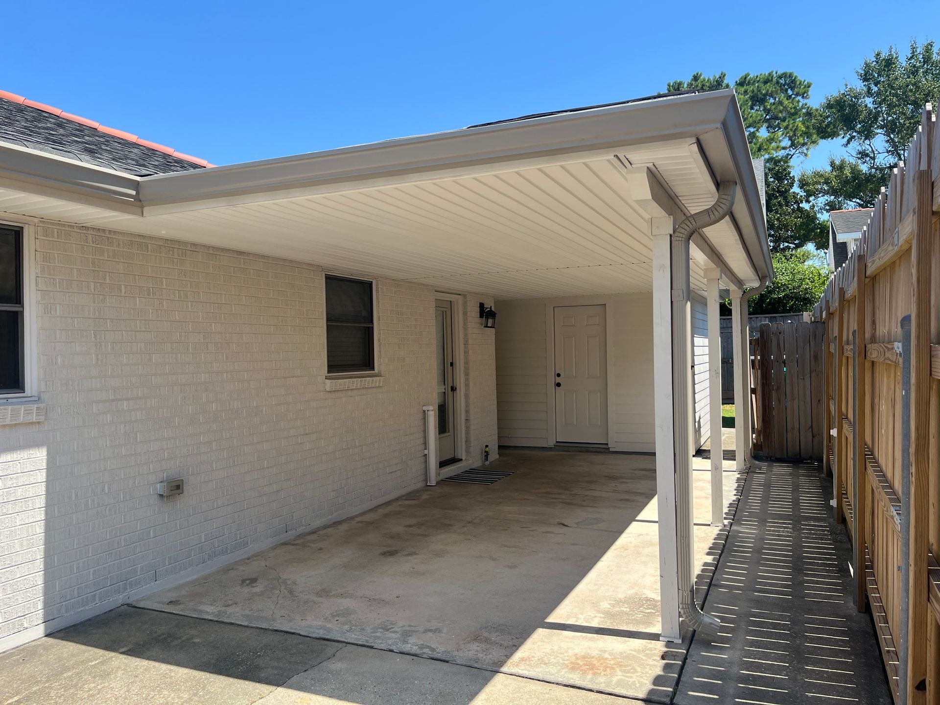 A beige brick house featuring a covered carport, white siding, and a wooden fence along the side on a sunny day.