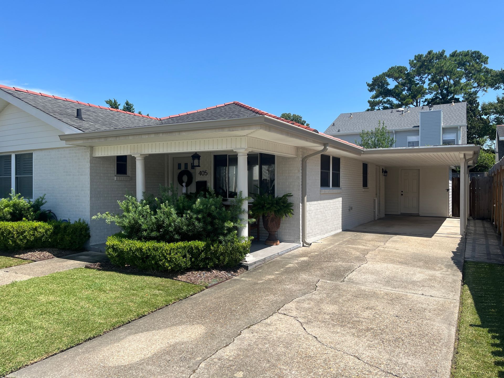 A single-story white brick house with a front porch, concrete driveway, and attached carport under a clear blue sky.