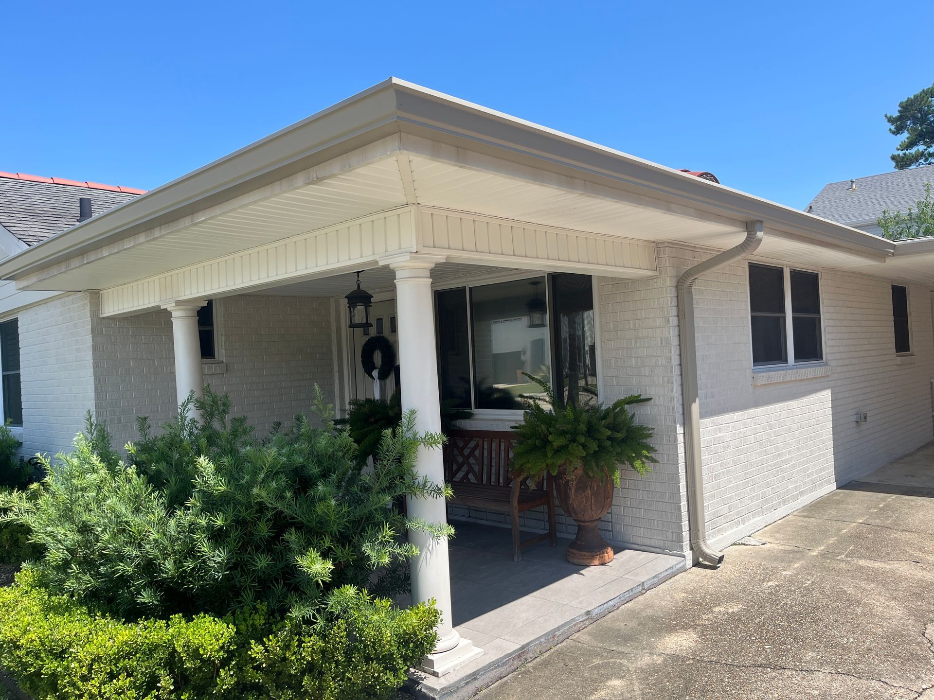 A covered white brick porch of a house with a concrete walkway, white columns, and lush green landscaping.
