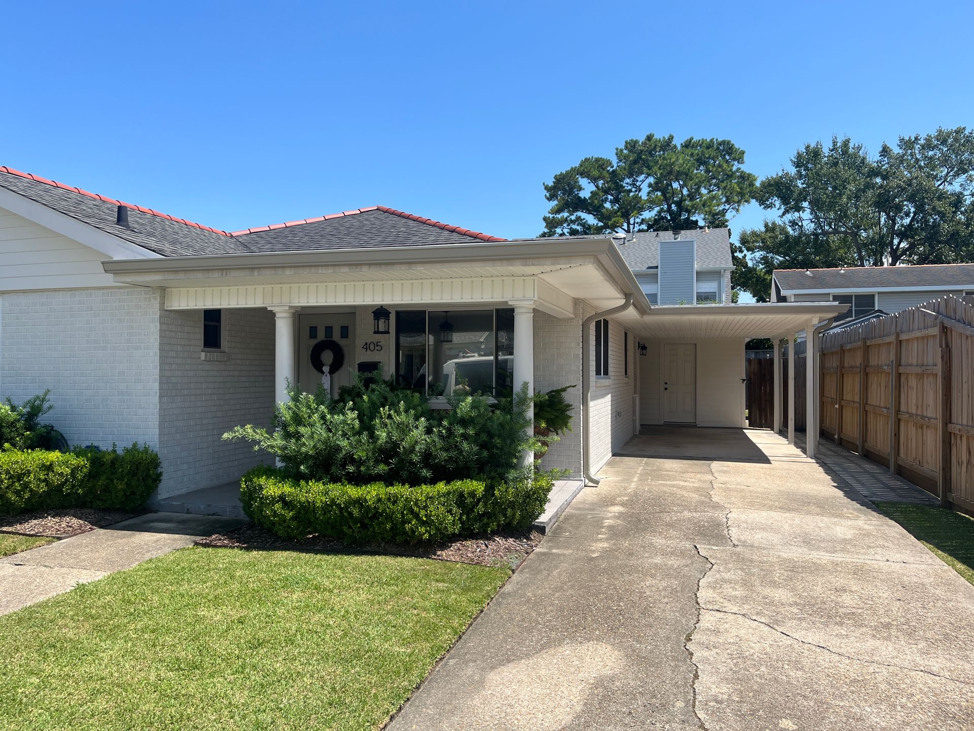 A single-story white brick house with a front porch, a green hedge, and a concrete driveway under a clear blue sky.
