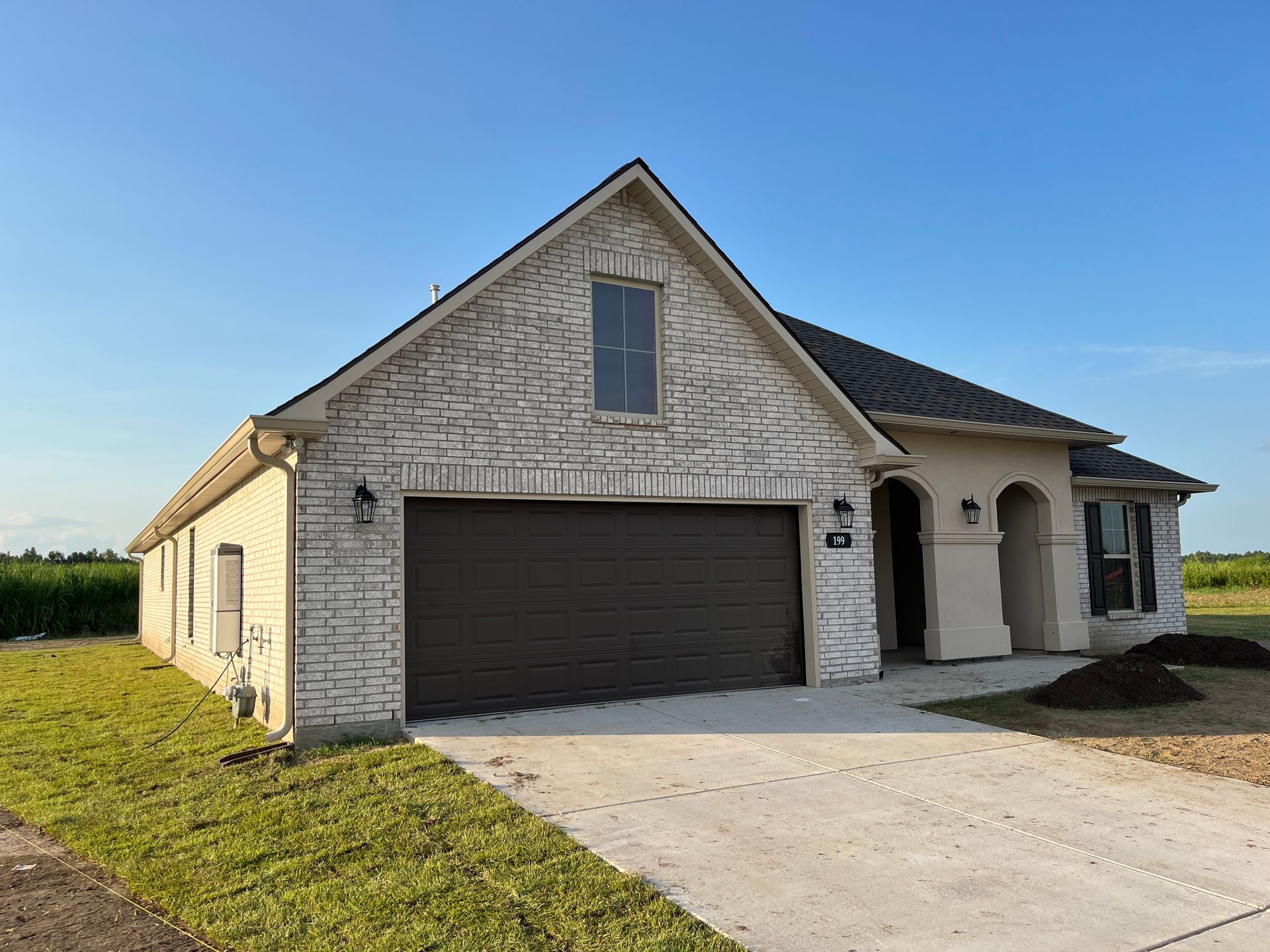A single-story suburban house with white-washed brick, a dark garage door, and a concrete driveway under a blue sky.