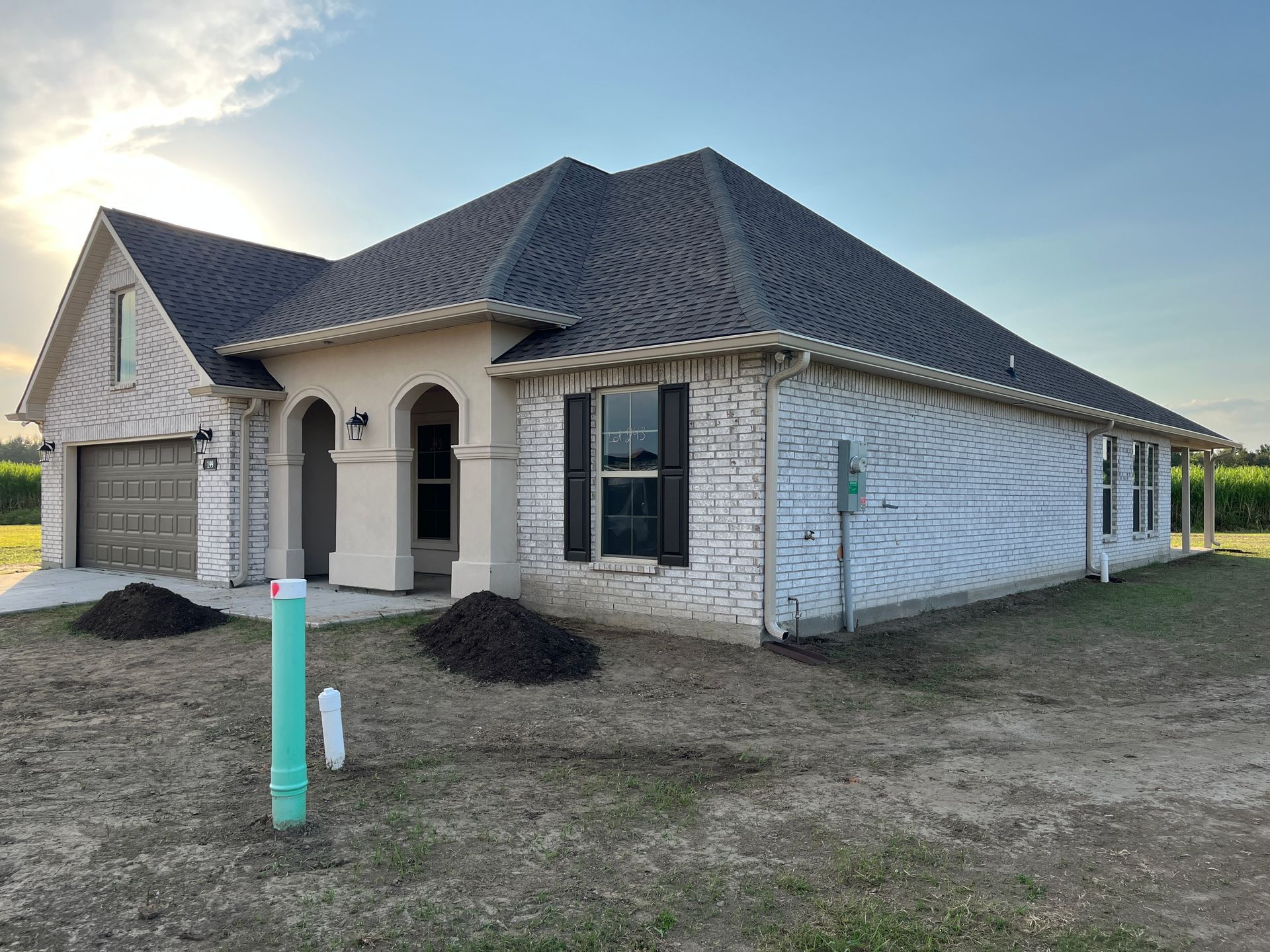 Single-story house with cream brick, tan trim, dark shutters, and a gray shingled roof under a clear blue sky.