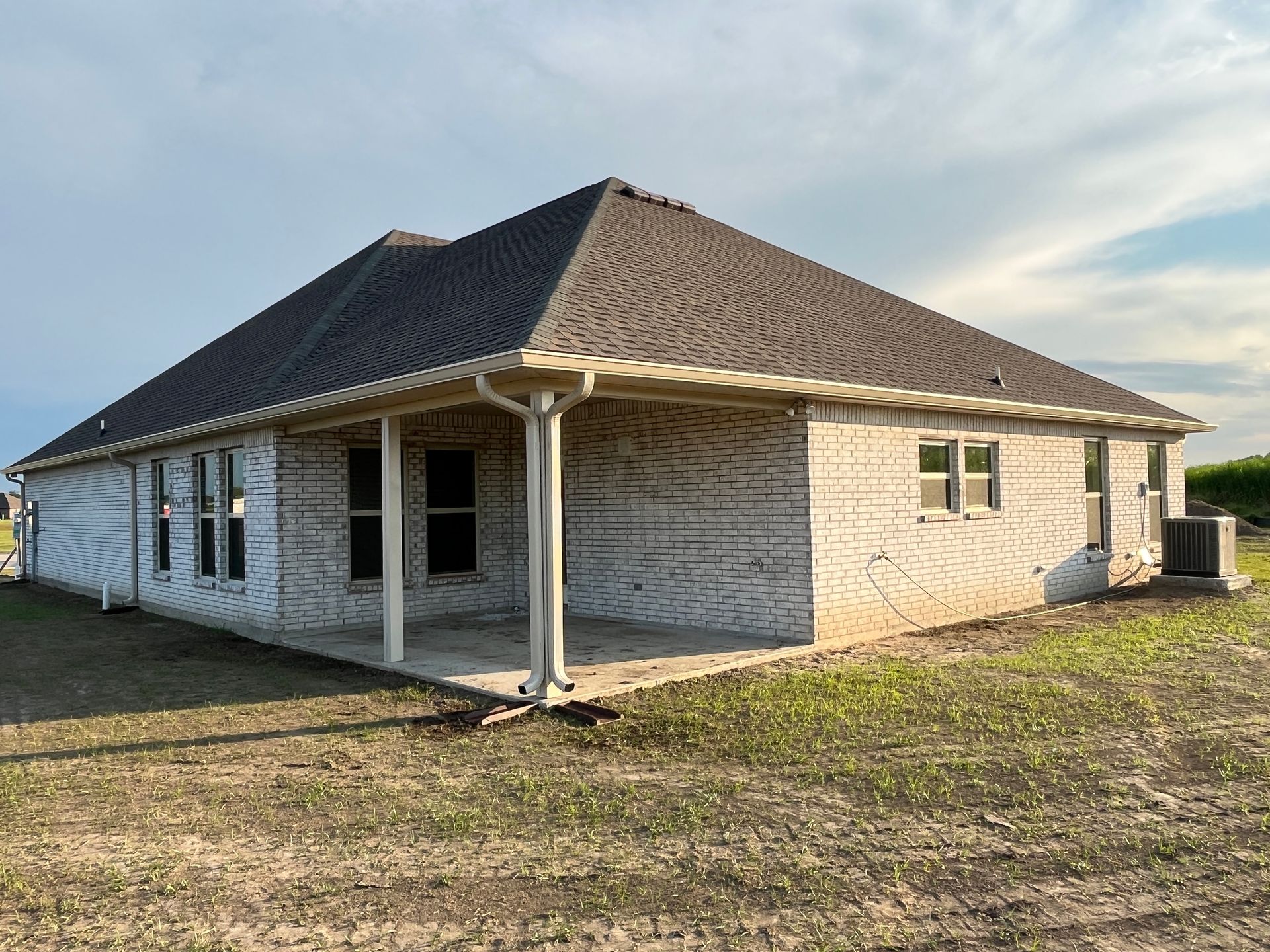 A white brick house under construction with a dark roof and a covered patio, set in an open, undeveloped yard.