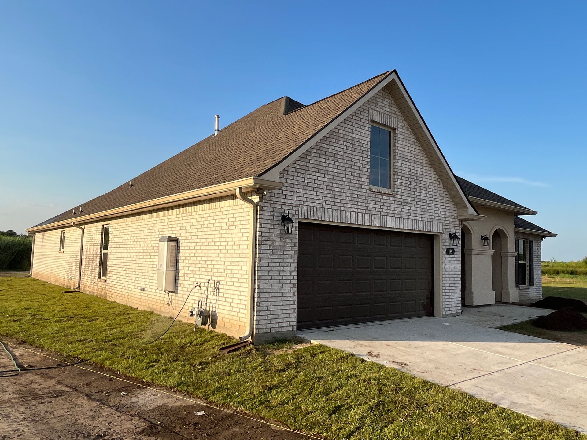 A single-story, light-colored brick house with a dark garage door, steep roofline, and concrete driveway under blue sky.