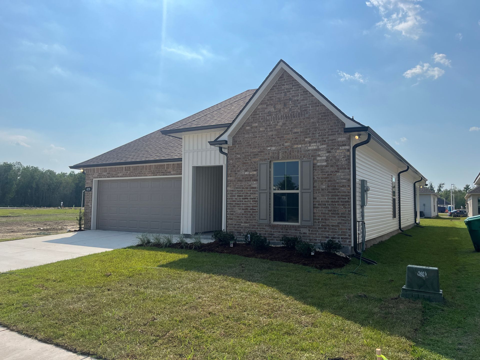 A tan and brown brick house with a white side, a gray garage door, and a grass lawn under a clear blue sky.