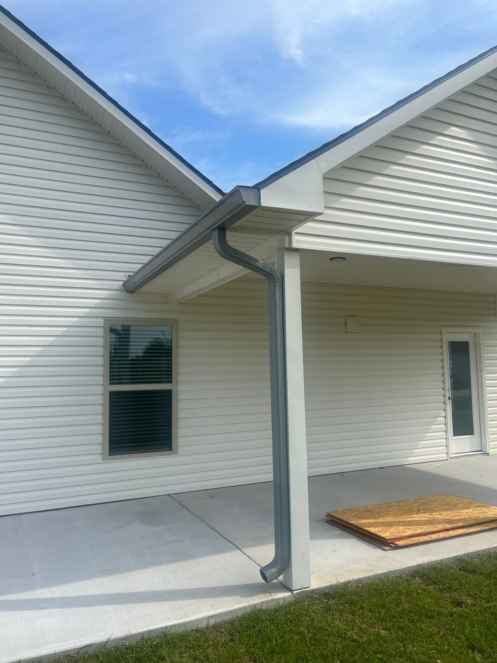 A corner of a white-sided house with a covered concrete patio, grey gutters, and a vertical downspout attached to a post.