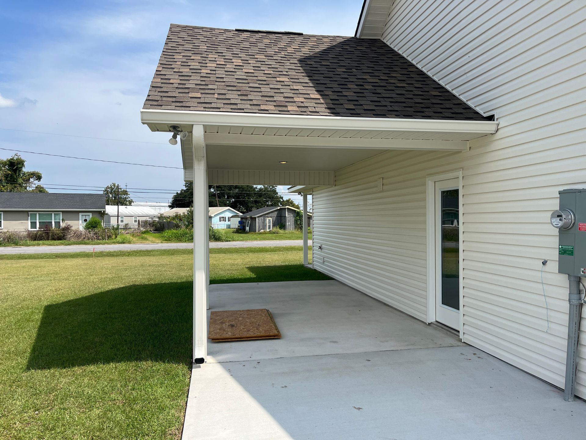 A concrete patio sheltered by a roof overhang attached to the side of a white-sided building with a door.