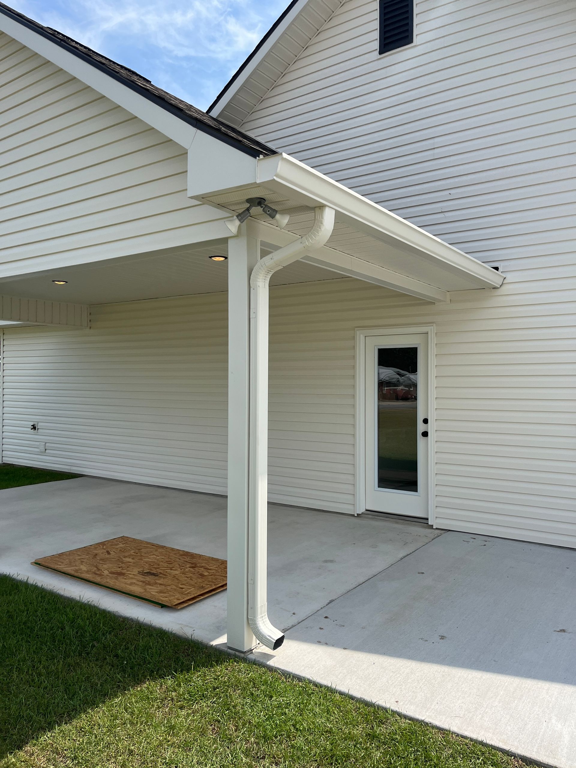 A white exterior house wall with a covered concrete patio, a doorway, and a vertical white gutter downspout.