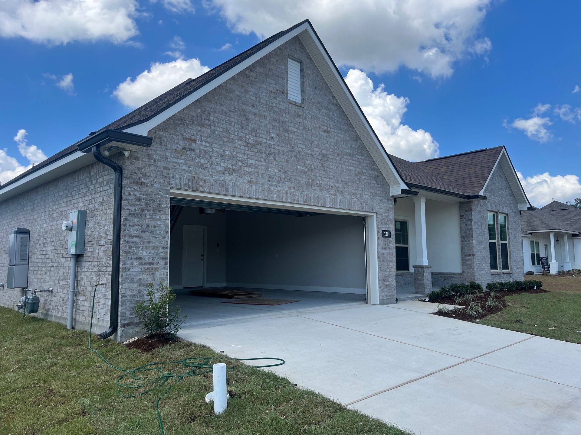 A newly constructed, single-story gray brick house with an open two-car garage and a concrete driveway under a blue sky.