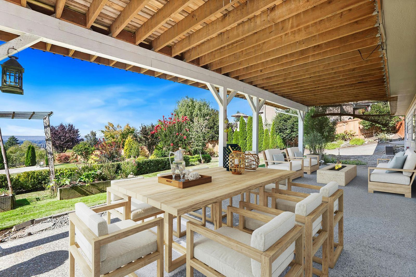 Outdoor patio with a long wooden table and chairs, overlooking a lush garden with a blue sky.