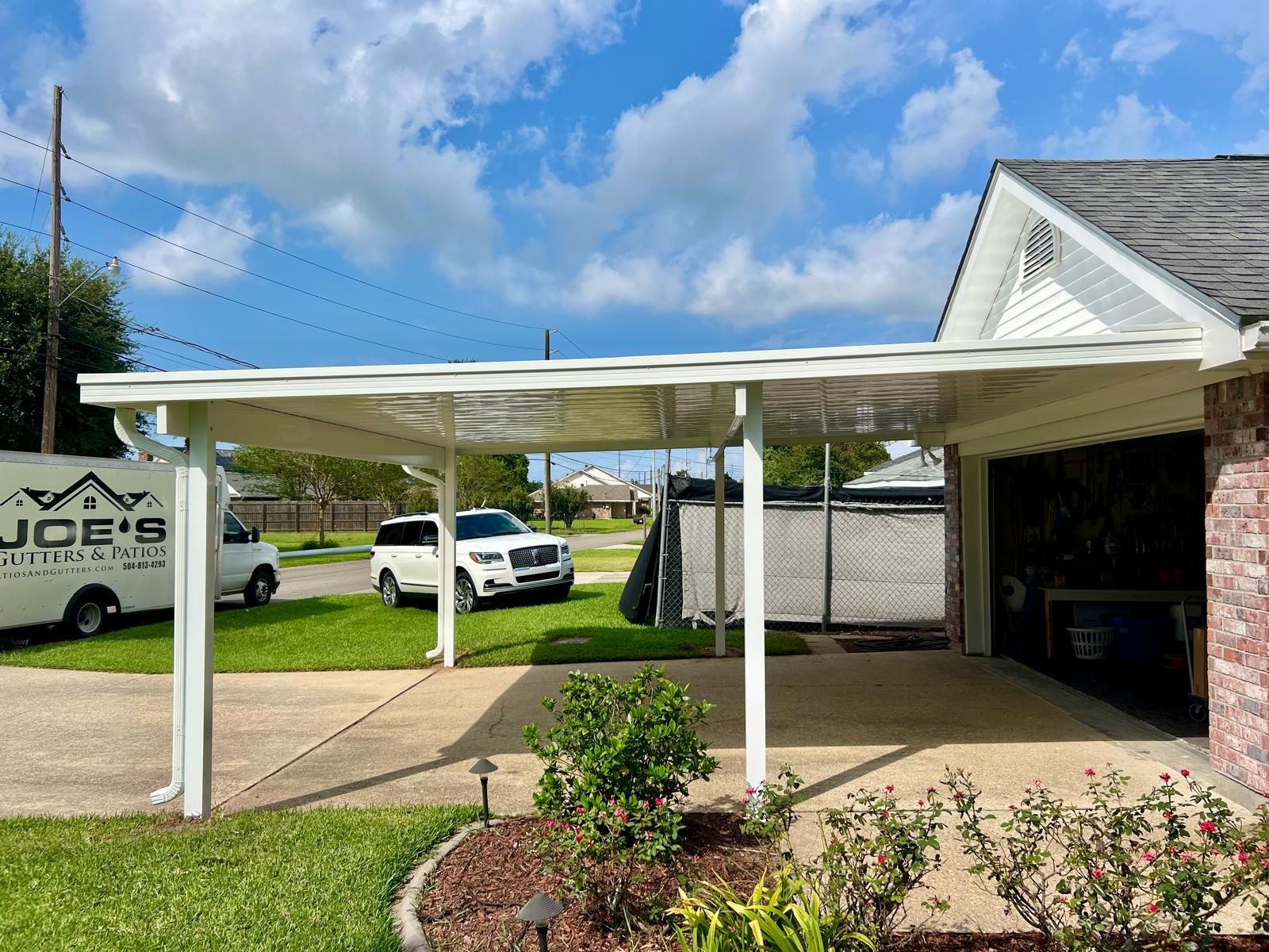 A white metal carport attached to a house, with a driveway, a parked white SUV, and a service van in the background.
