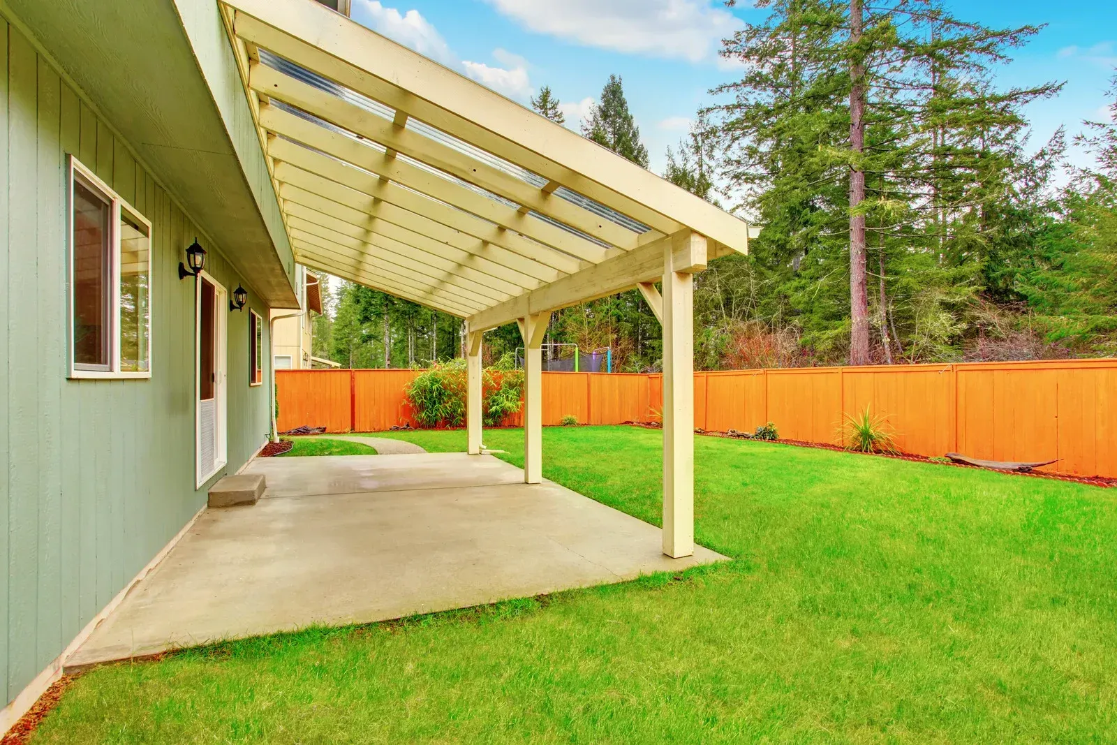 Green house with a concrete patio and pergola. Orange fence and green lawn.