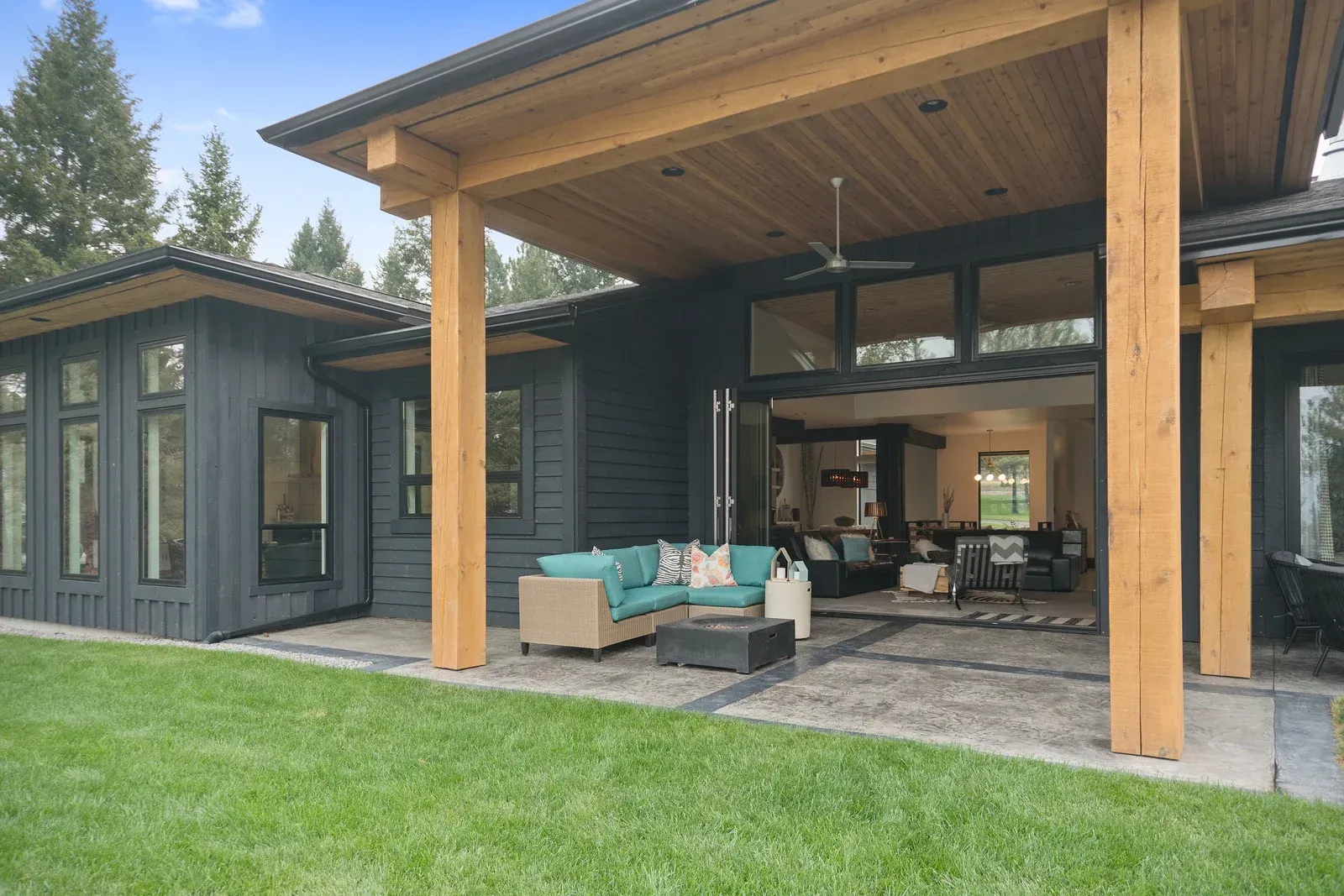 Black house with wooden columns and an outdoor patio with furniture, next to green lawn.