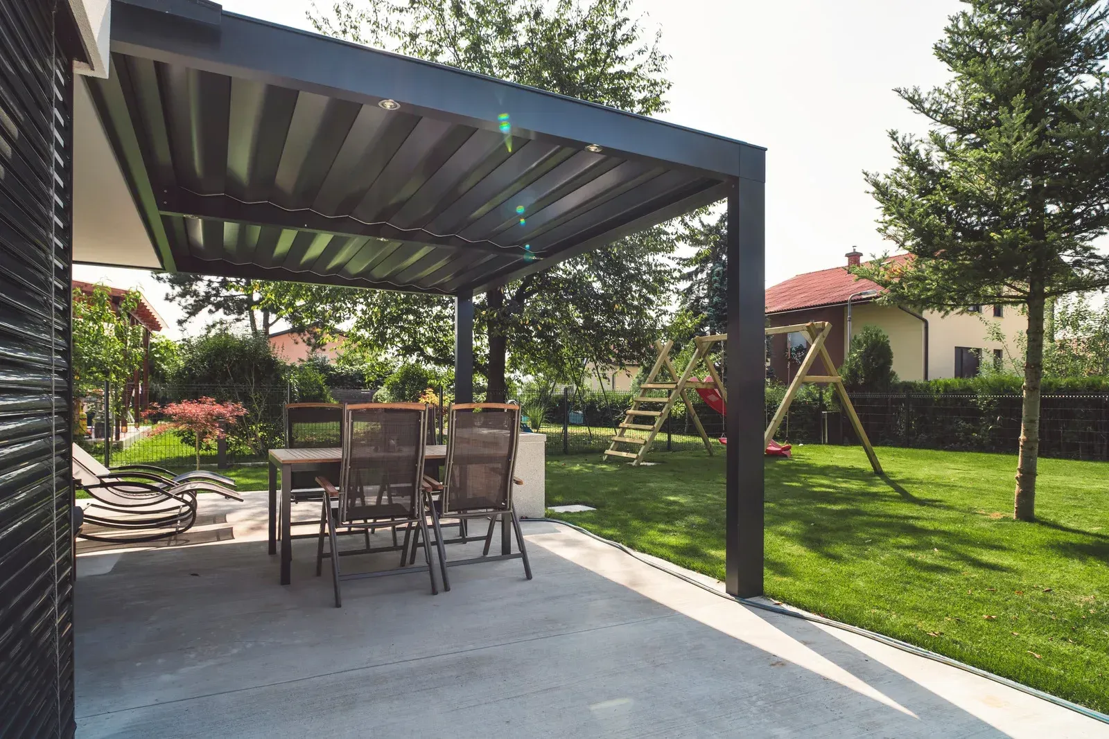 A gray patio with a pergola, table, chairs, and a backyard with swing set and trees.
