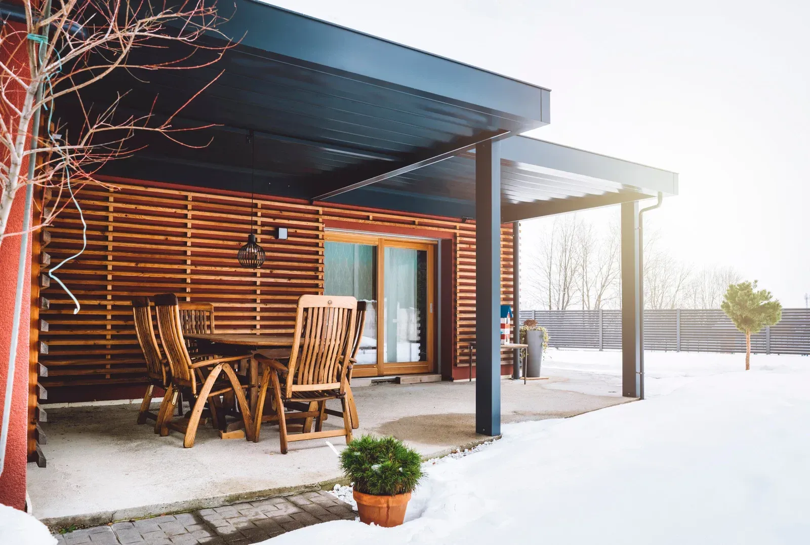 A wooden patio with table and chairs, covered by a dark roof, in a snowy setting.