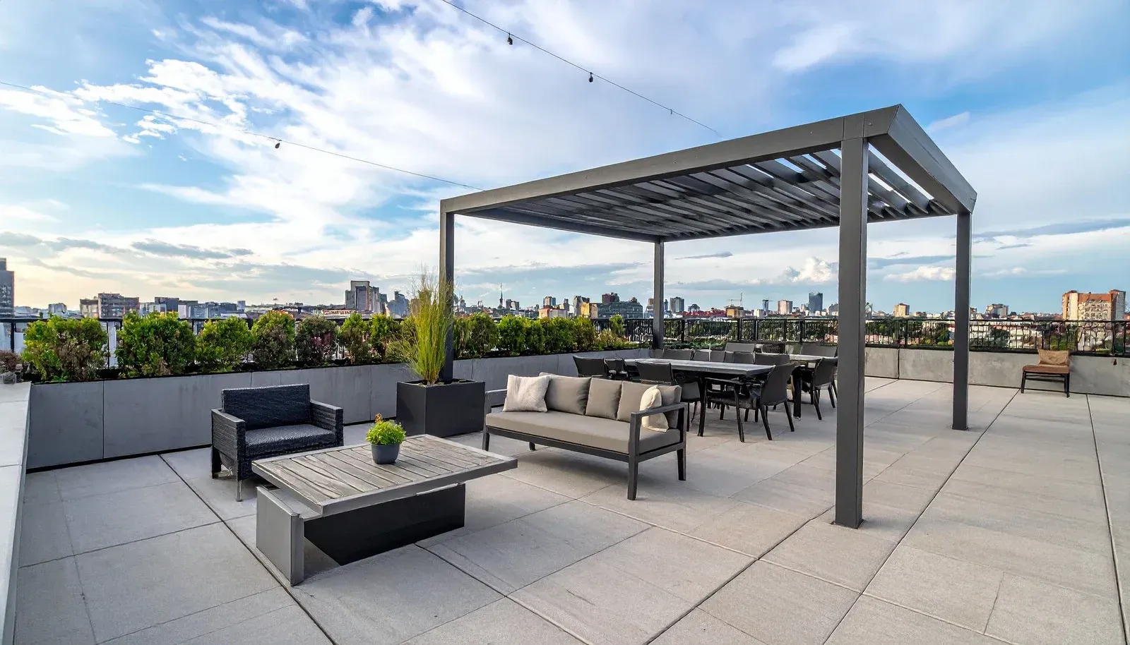 Rooftop patio with seating, dining table, and pergola, overlooking city skyline under a blue sky.