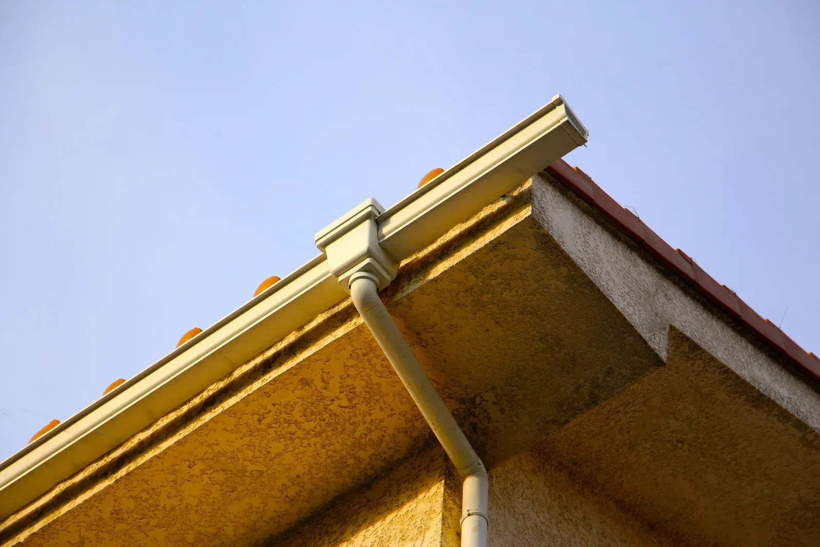 Corner of a building with light-colored gutters, a downspout, and a reddish tiled roof against a blue sky.