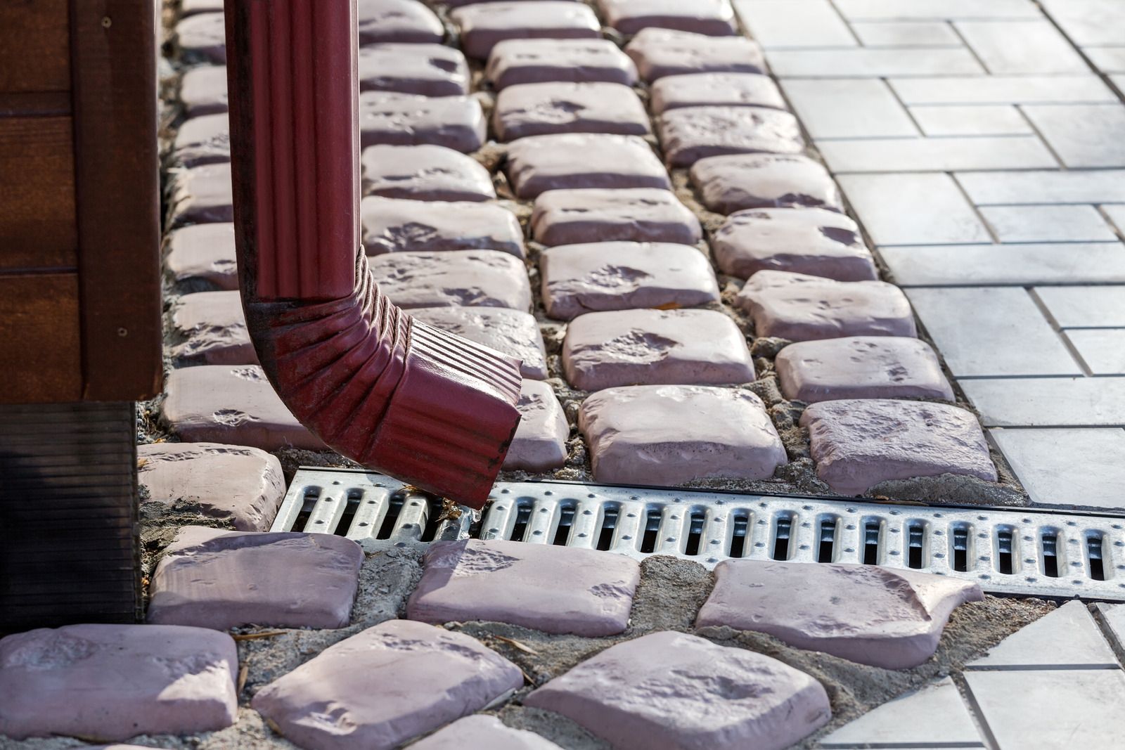 Red rain gutter directs water to a metal grate in brick paved walkway.