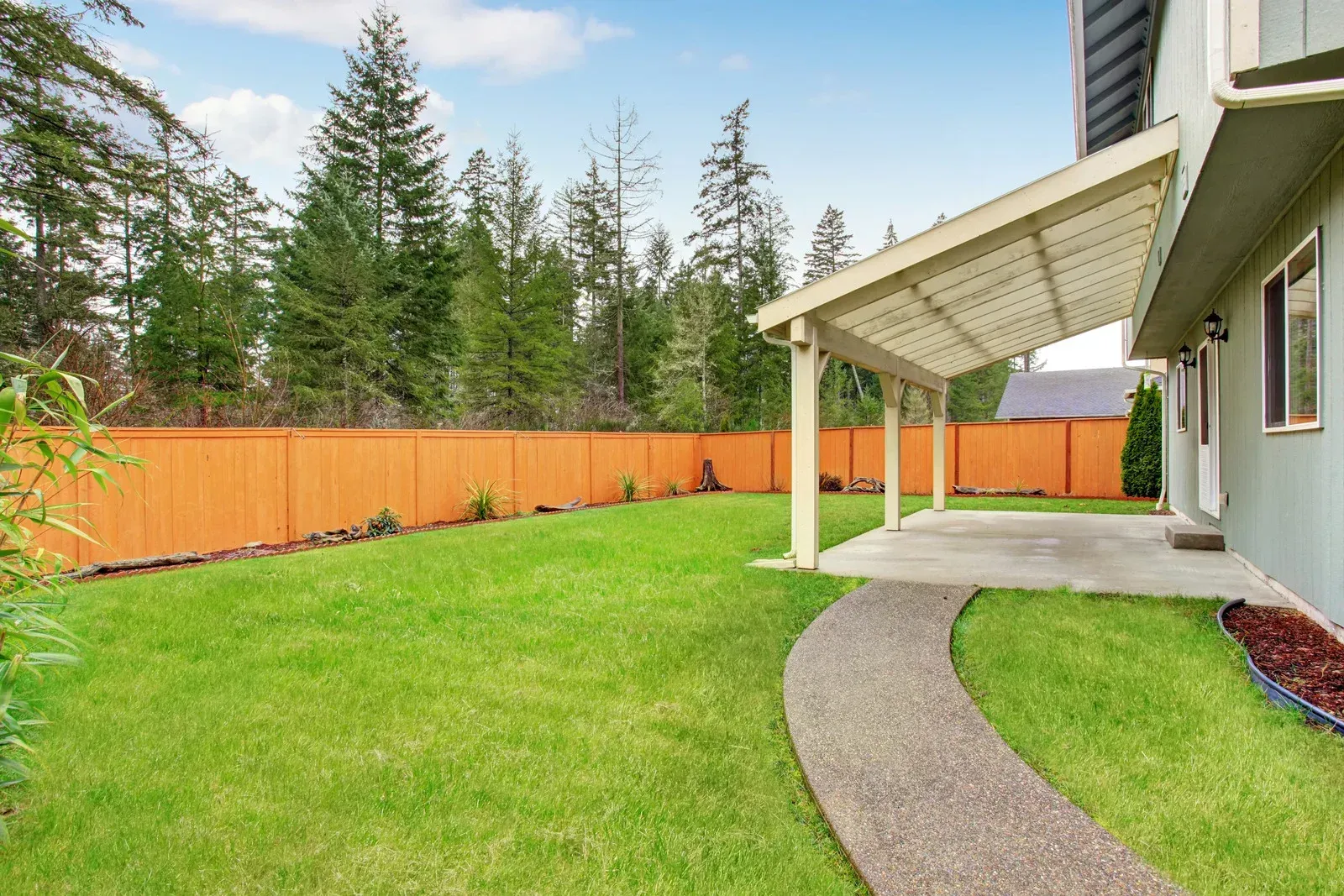 A green lawn with a curved stone path leads to a covered patio next to a house with a wooden fence and trees.