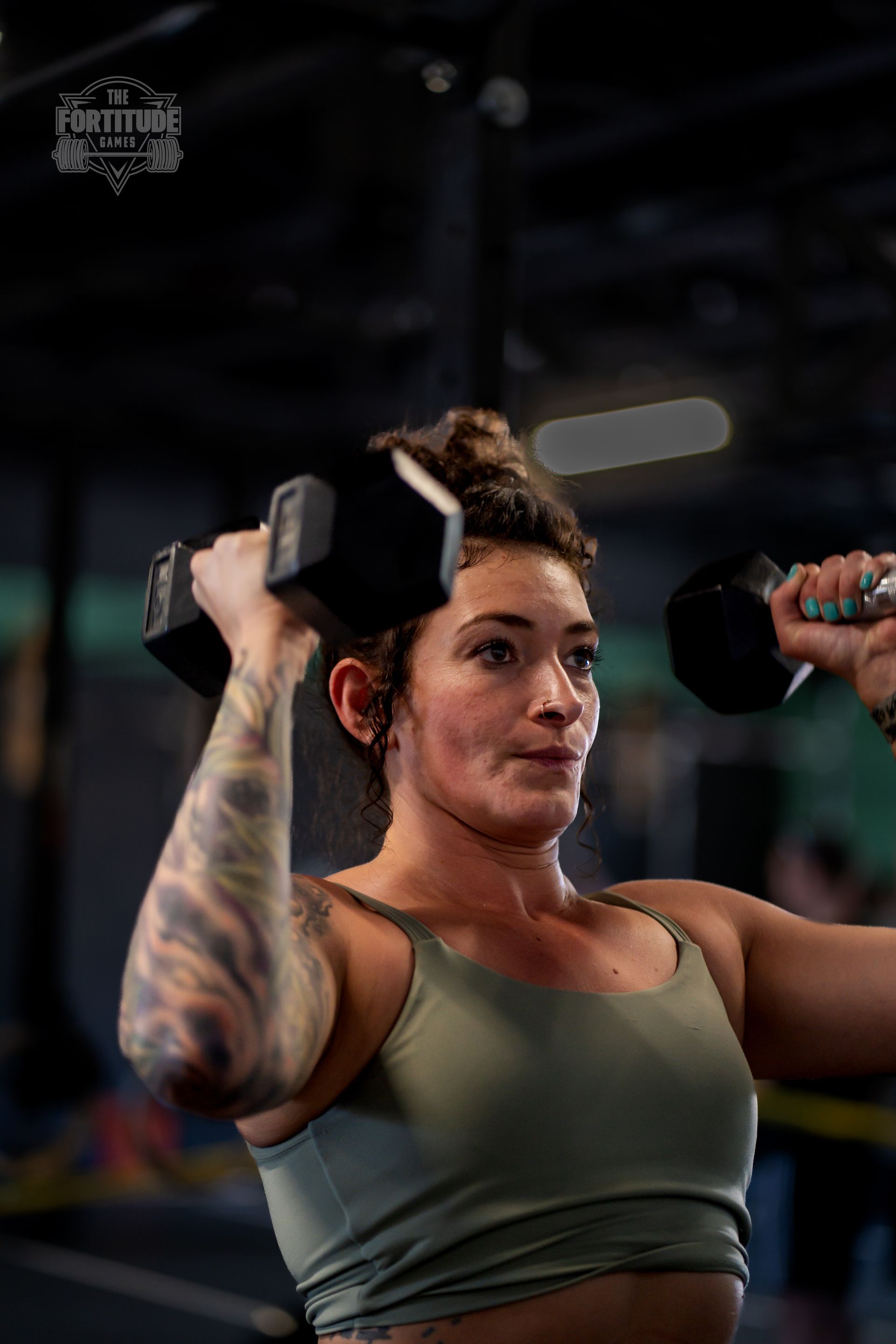 Woman lifting dumbbells overhead in a CrossFit gym in Lakewood, CO, wearing a sports bra, with arm tattoo.