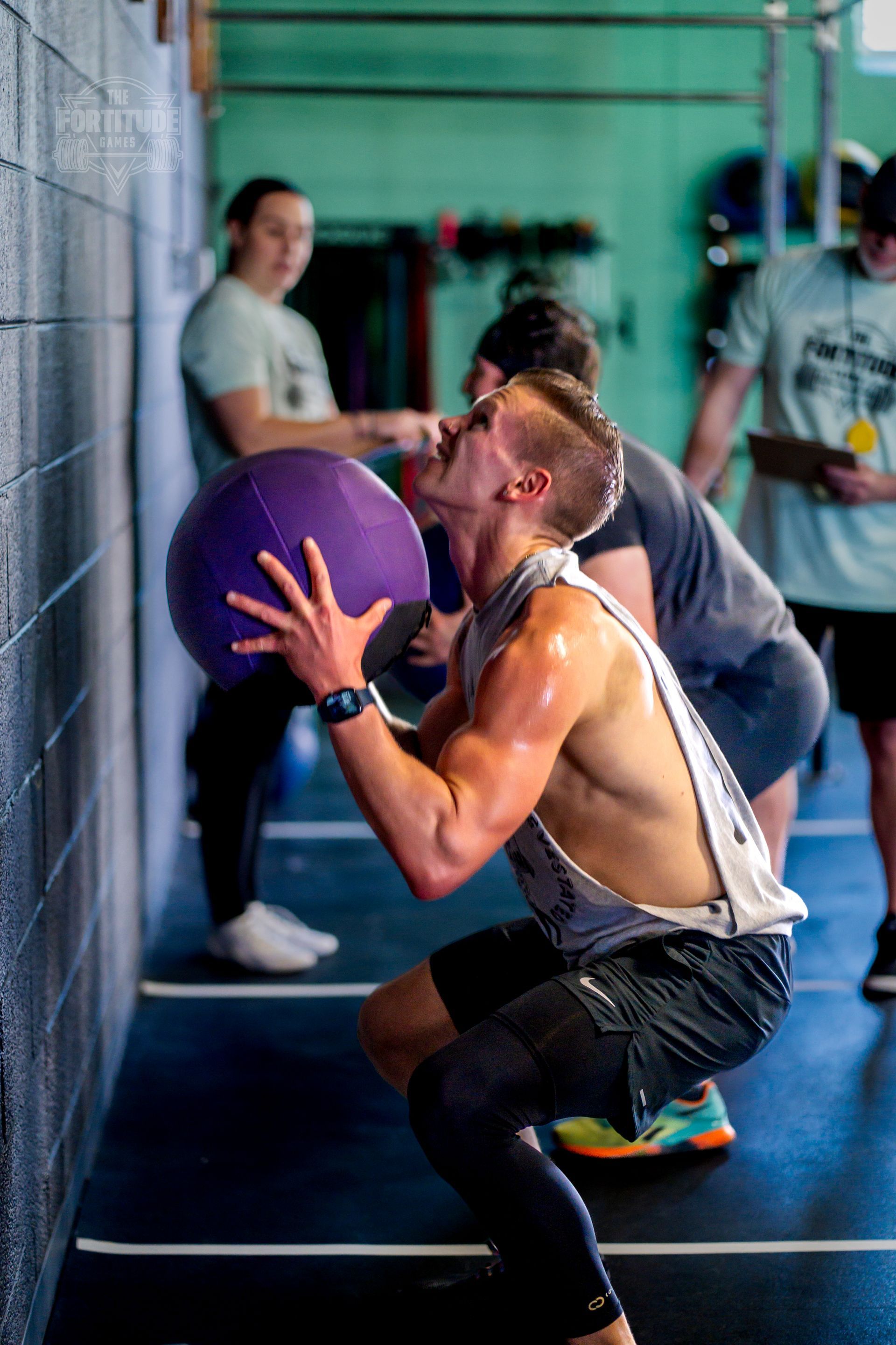 Man doing wall ball squat, sweating, at a gym in Lakewood, CO. Others watch. Purple ball, blue wall.