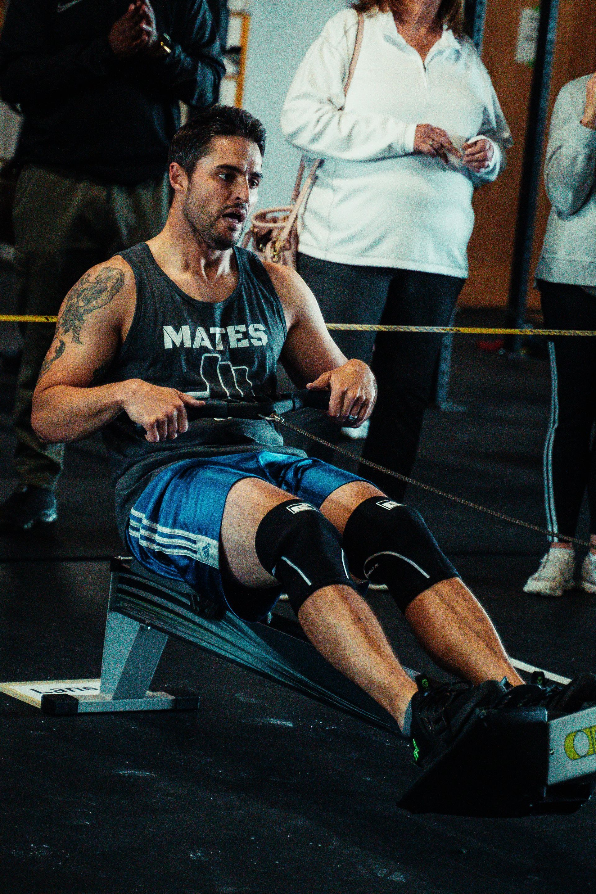 Man rowing in a Lakewood, CO gym, wearing a tank top, blue shorts, and knee pads. People watching in the background.