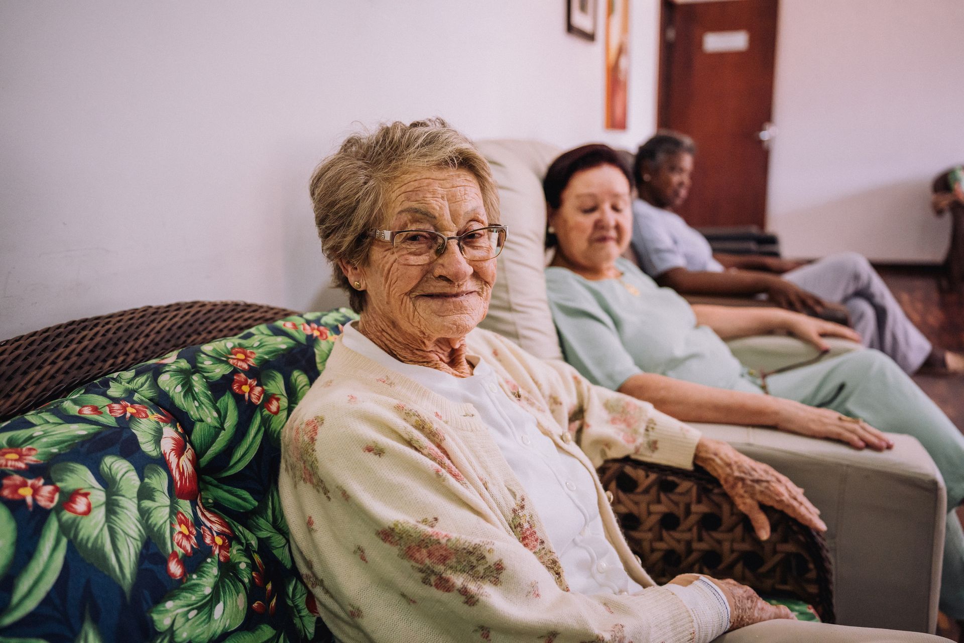 Three elderly people sit on a floral couch in a cozy communal area.