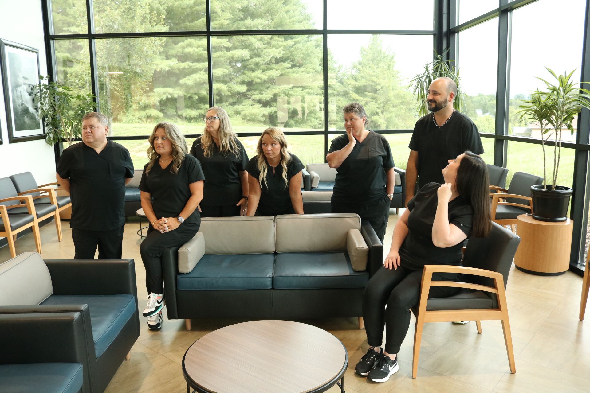A group of seven people in black standing and sitting in a modern waiting room with large windows.