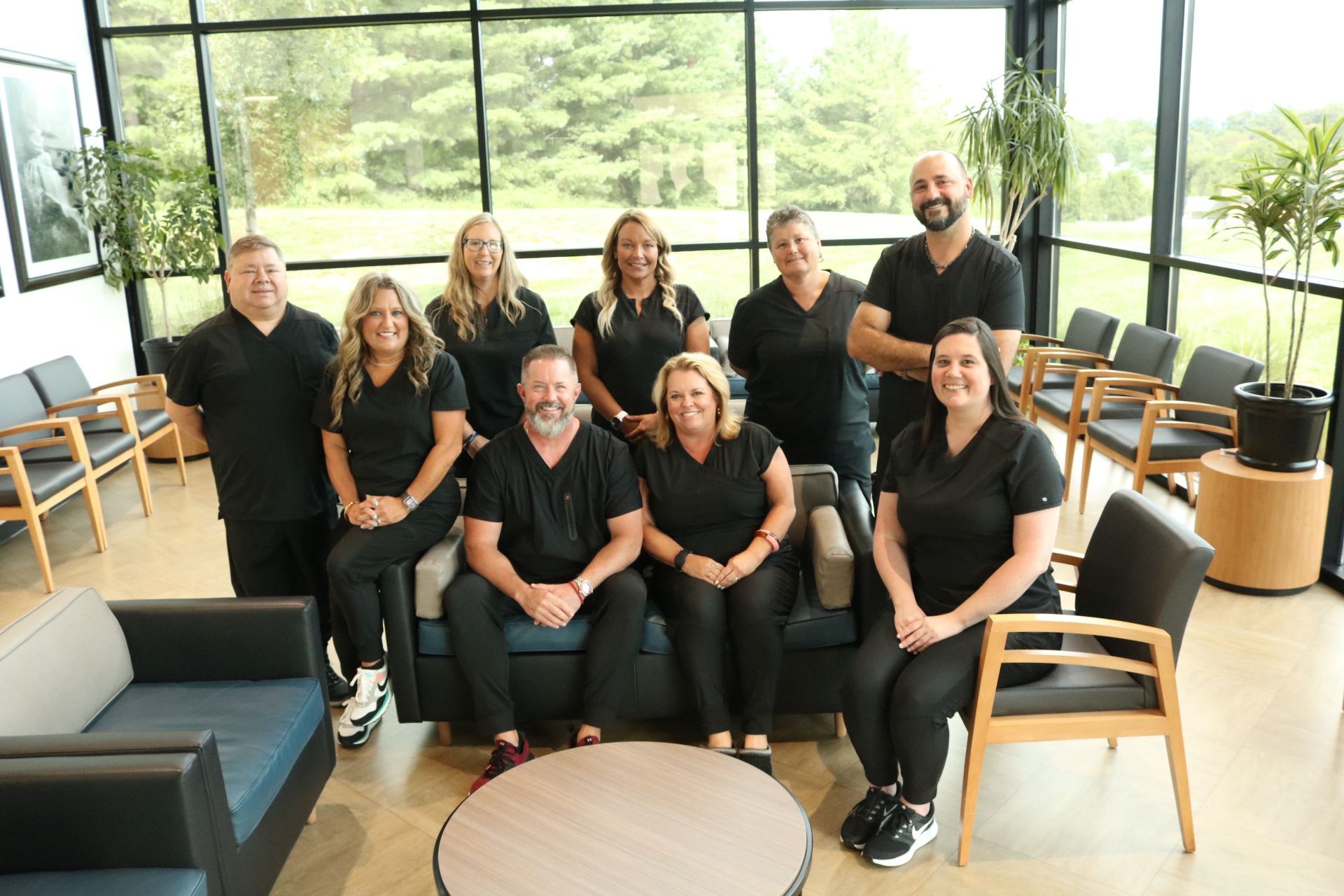 Dental clinic staff posing together: nine in black scrubs, sitting and standing indoors with large windows.