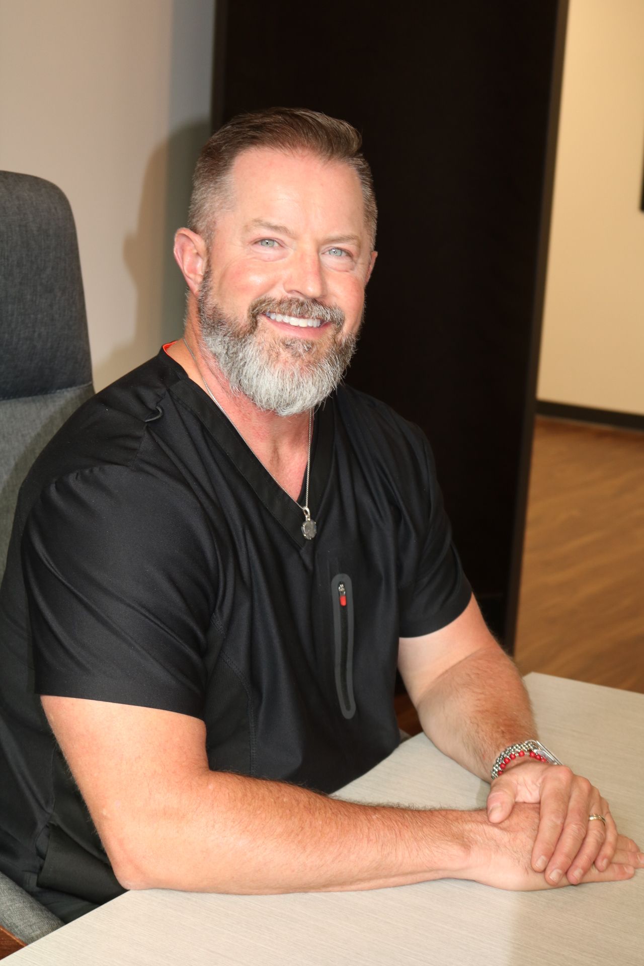 Man with gray beard smiles, wearing black scrubs. Leans on a table in an office setting.