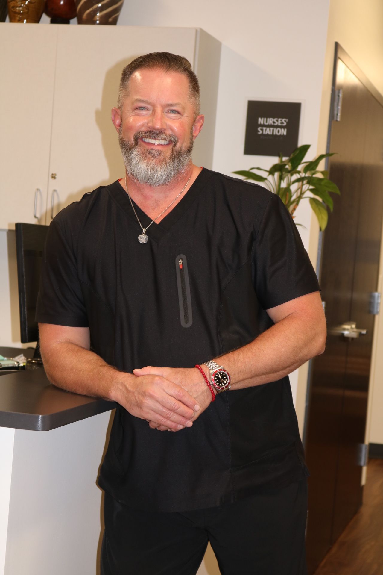 Man with beard in black scrubs, smiles in a modern clinic setting.