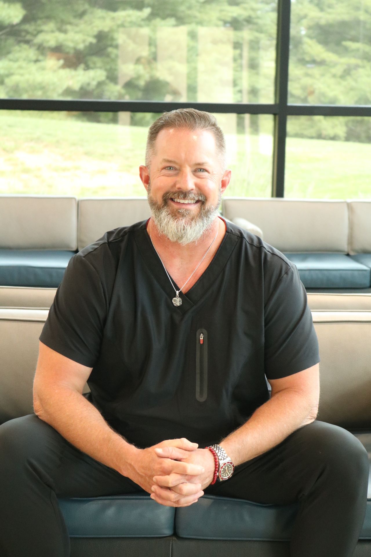 Man with gray beard, in black scrubs, smiling, seated on blue couch.