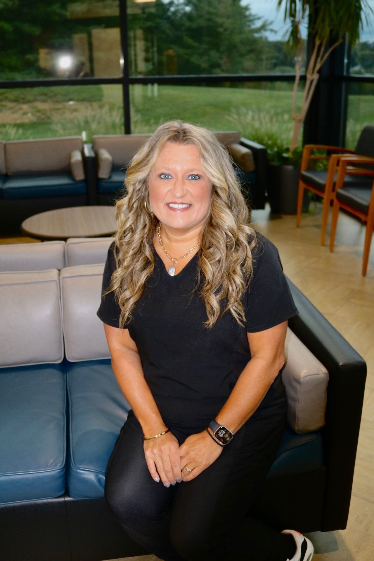 Woman with blonde hair smiles, wearing black scrubs, sitting on a blue couch in a modern office.