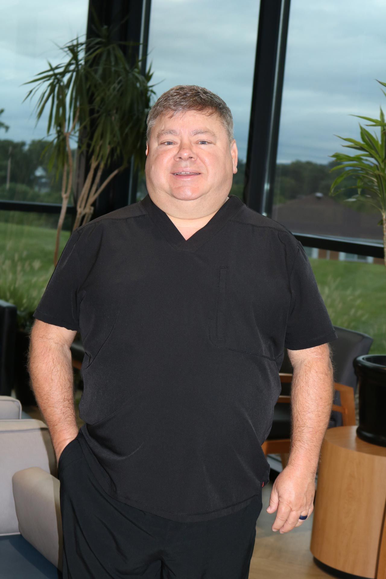 Man in black scrubs poses by a window, hands in pockets, smiling.